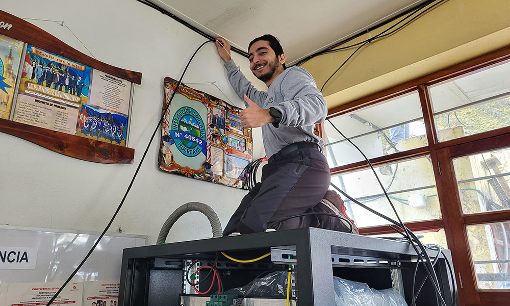 A man kneels on a large box containing electronics while holding a wire up to the ceiling. he looks back at the camera and smiles and gives a thumbs up.
