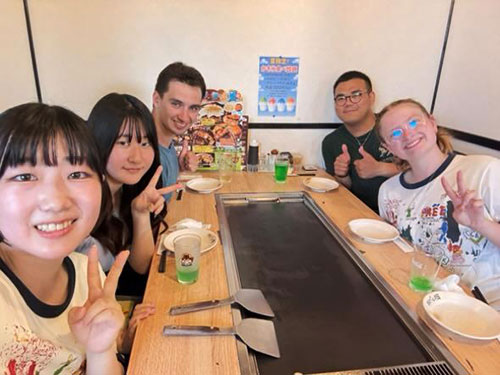 Five people seated around a table with a built-in grill at a restaurant, smiling and making peace signs. Plates, drinks—including green beverages—and spatulas are on the table. A colorful menu is visible on the wall behind them.