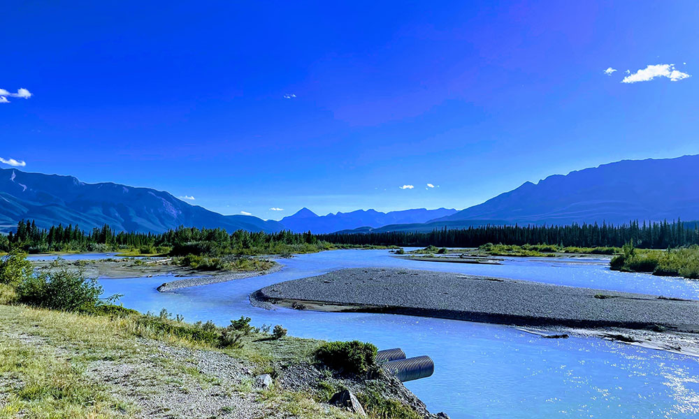 A wide shot of a bright blue river meandering through a valley, with mountains in the background under a clear blue sky with a few white clouds.