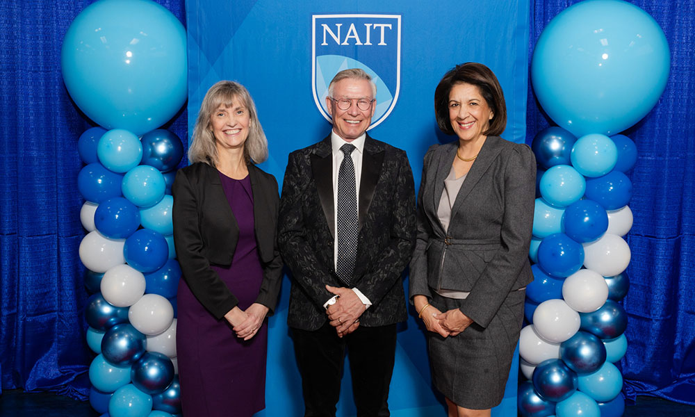 three women pose for a photo with a man standing between them. they are wearing business and formal attire. behind them is a blue background and blue balloons.