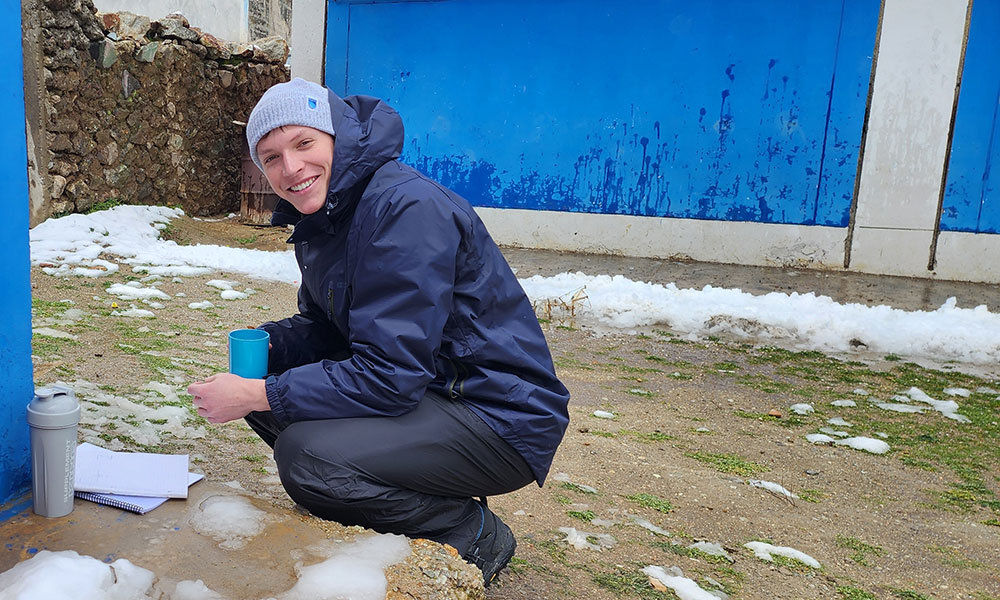 a man in a blue jacket and light blue toque crouches and smiles for the camera. he is holding a blue mug