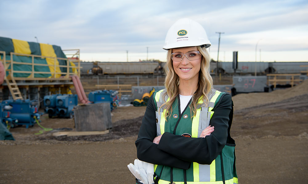 Person wearing a hard hat and safety vest stands with arms crossed at an active construction site with equipment and railcars in the background.