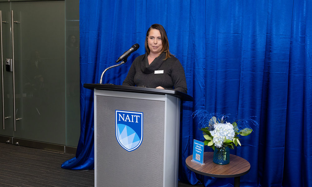 A person stands at a podium with a microphone in front of a blue curtain. The podium displays the NAIT logo, and a small table beside it holds a vase of white flowers and a blue booklet.