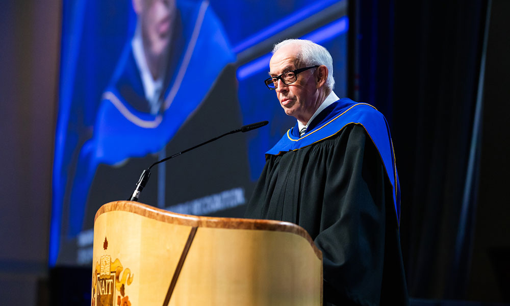 Person in academic regalia delivering a speech at a podium with the NAIT logo. A large screen behind displays an enlarged image of the speaker."