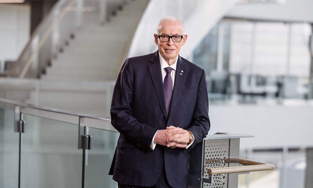 a older man wearing black-rimmed glasses and a dark suit jacket with a pale shirt and purple tie poses for a photo in a modern-looking building