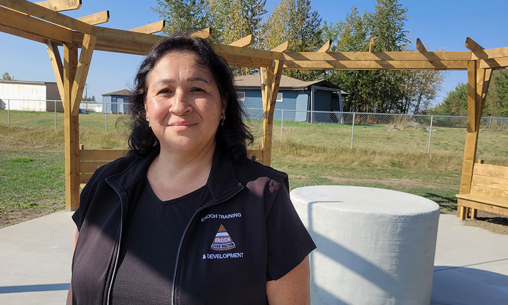 A person wearing a black vest with an “Enoch Training & Development” logo stands outdoors under a wooden pergola structure, with grass, trees, and buildings visible in the background.