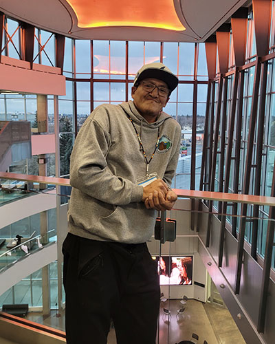 A person in a grey hoodie and cap leans casually on a railing inside a modern glass-walled building with a high ceiling and warm lighting.
