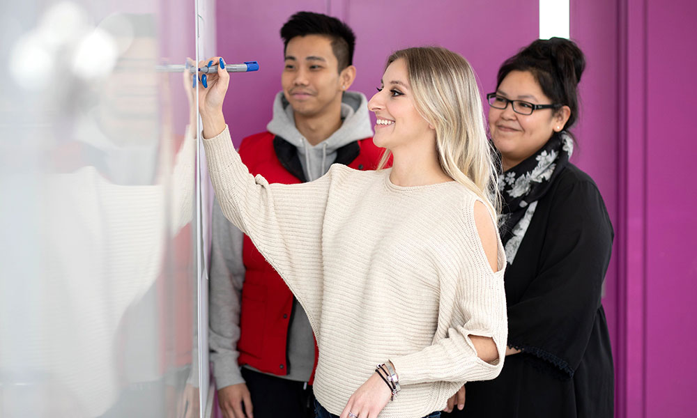 woman in white shirt drawing on white board with blue pen, two students watch her and smile