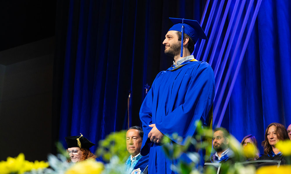 Person in a blue graduation gown and cap standing on a stage, with blue curtains in the background. Several others in graduation attire are seated behind, and yellow flowers are visible in the foreground."