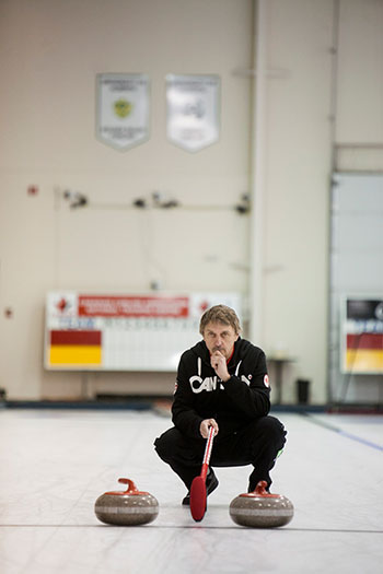 A person crouching on an indoor curling rink with a broom, preparing a move with two curling stones in front, surrounded by curling-related banners.