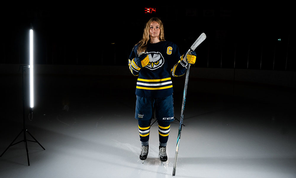 A hockey player in a dark blue uniform with yellow and white stripes stands on an ice rink holding a stick upright. Bright vertical lighting illuminates the scene against a dark background with a faint red scoreboard overhead.