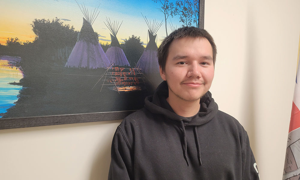 A person wearing a black hoodie stands indoors beside a wall with a colorful painting of tipis at sunset and part of a flag visible on the right.