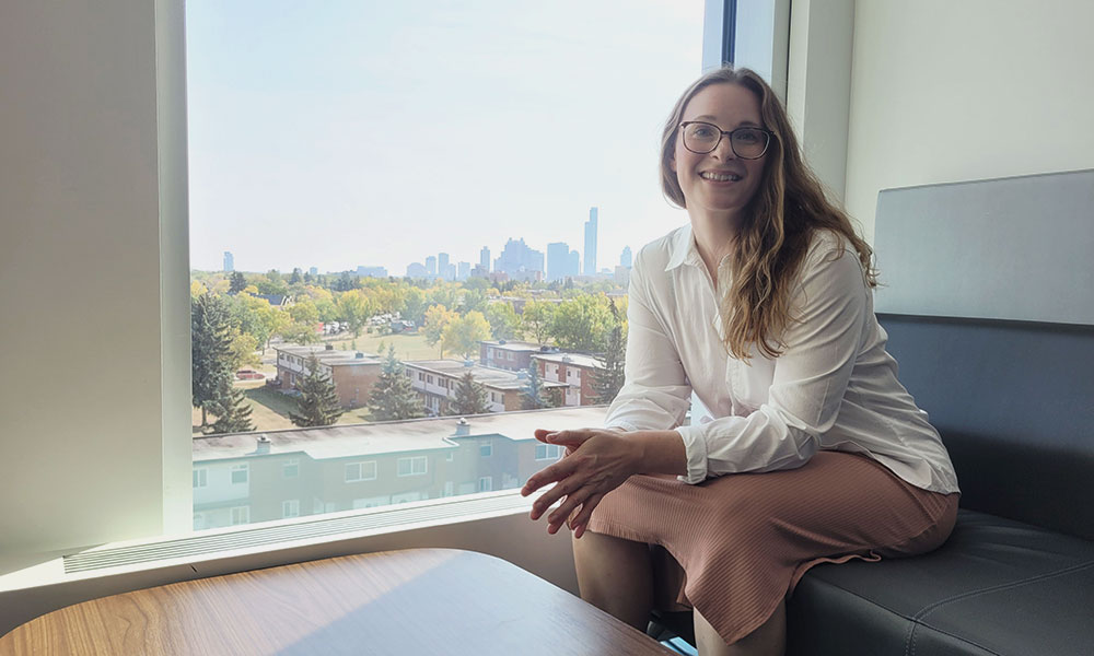 Person wearing glasses, a white shirt, and a light-colored skirt sitting on a bench near a large window with an urban skyline and trees visible in the background.