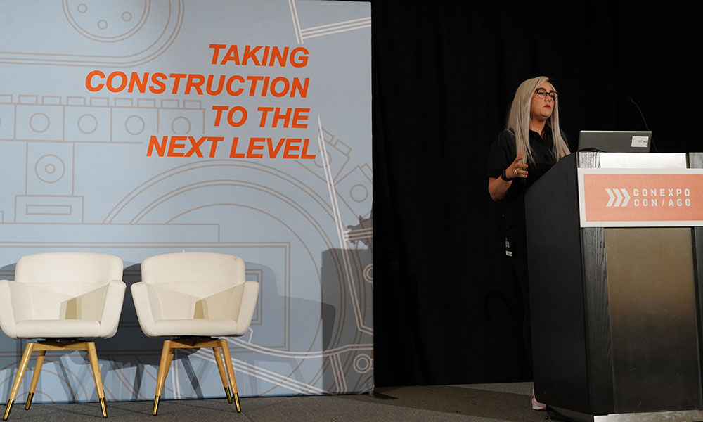 Speaker presents at a construction conference from a podium beside a stage backdrop reading “Taking Construction to the Next Level.”
