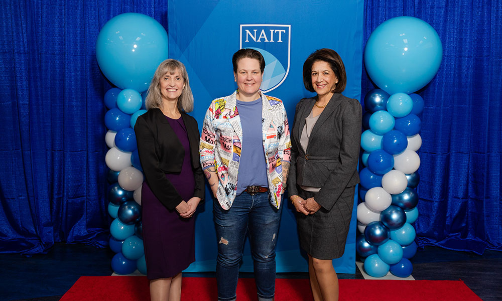 three women pose for a photo. they are wearing business and formal attire. behind them is a blue background and blue balloons.