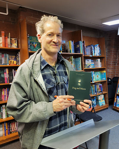 a man wearing a grey hoodie and a blue flannel shirt smiles and holds a copy of a green book called Dog and Moon