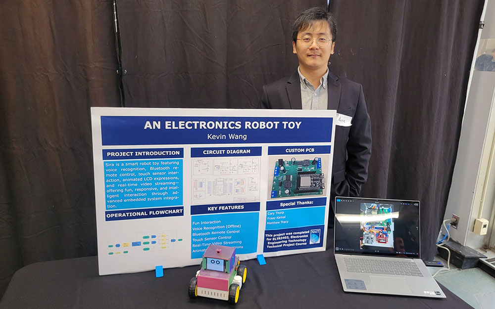 a young man in a dark blazer and pale blue shirt stands at a table with a project poster and a robot that looks like a small toy truck