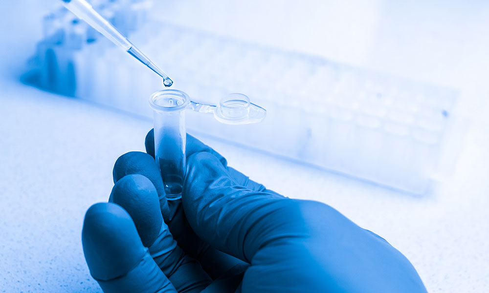 A gloved hand holds a small plastic test tube while a dropper releases a droplet of liquid into it. In the background, a rack holds multiple similar test tubes.