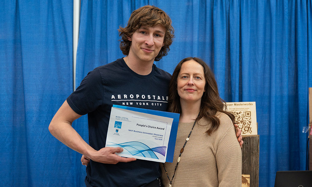 a man in a black shirt and holding a certificate stands with a woman in a brown sweater for a picture