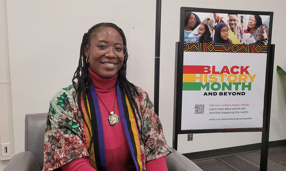 A person seated in a chair wearing a patterned shawl over a red top and multicoloured scarf, positioned beside a “Black History Month and Beyond” sign.