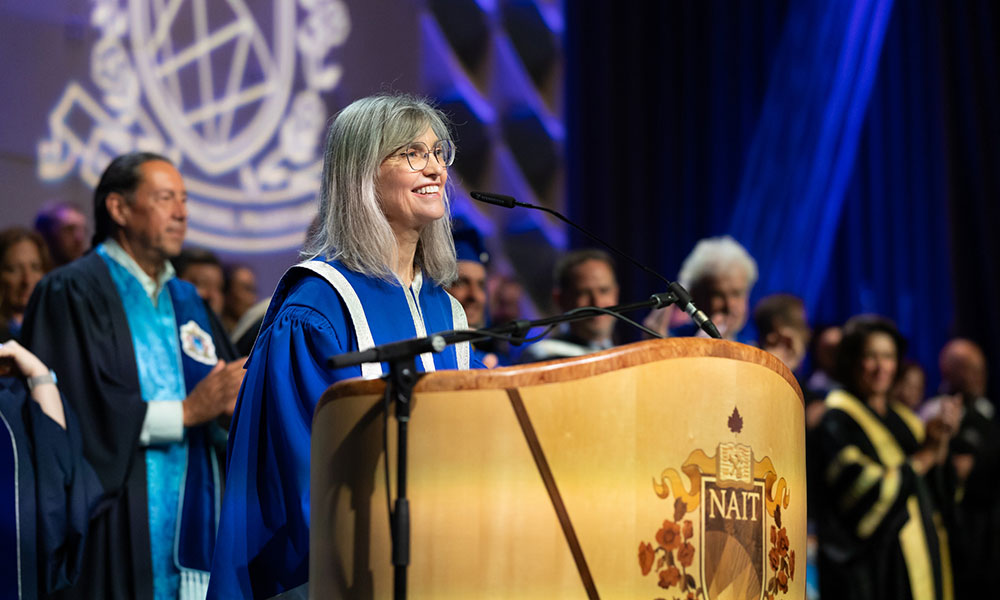 Person with gray hair speaking at a podium with the NAIT emblem, during a graduation or academic event. Several individuals in academic regalia stand behind, with a large emblem and blue lighting in the background.