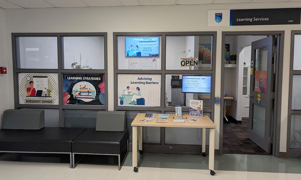 Interior view of a Learning Services area in an educational institution, featuring posters for 'Assessment Services,' 'Learning Strategies,' and 'Advising Learning Barriers.' A table with brochures, two black benches, and a door labeled 'Learning Services E105' are visible.