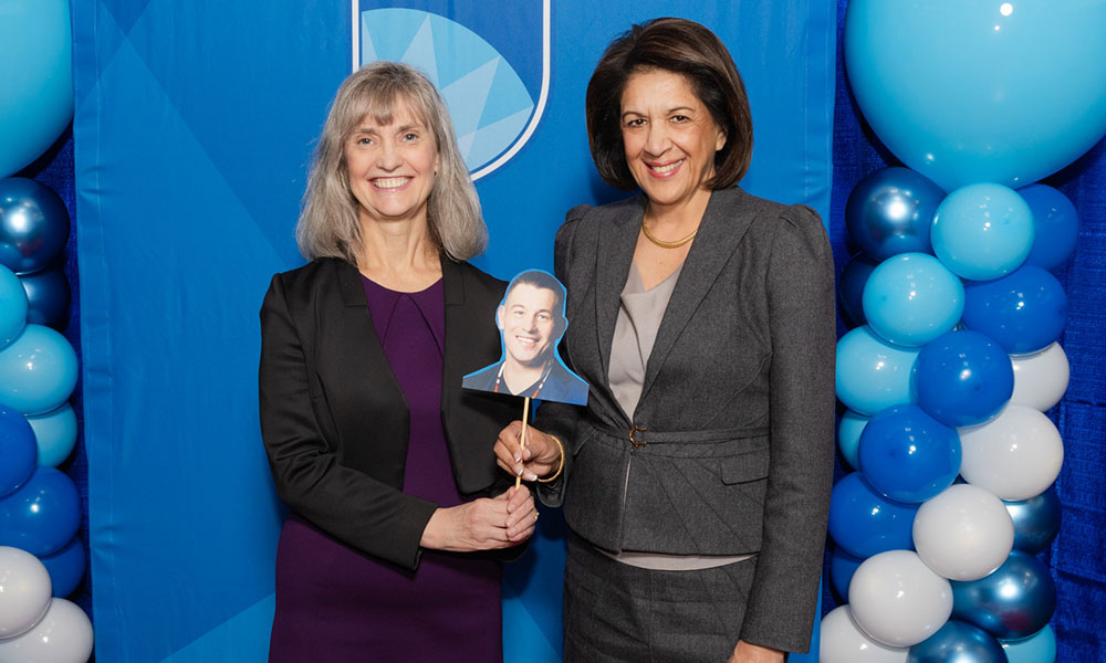 two women pose a photo holding a picture of a man's face on a stick. there is a blue background behind them