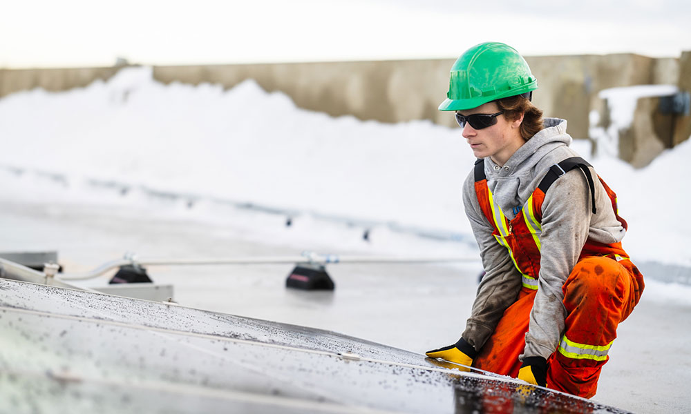a man in sunglasses, a green hard hat and orange coveralls kneels to put a solar panel in place on a snow-covered rooftop.