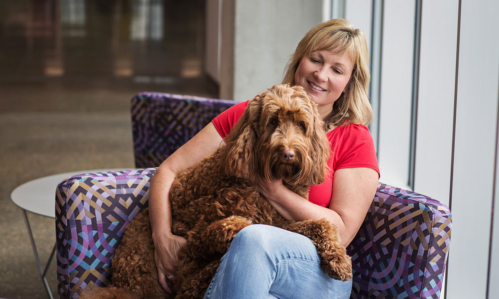 a woman in a red shirt holds a medium-sized shaggy brown dog as they sit in a mostly purple arm chair