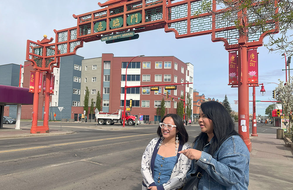 two women stand at a street corner. above them over the street is a red, metal chinese-style gate with the words "gate of happy arrival"