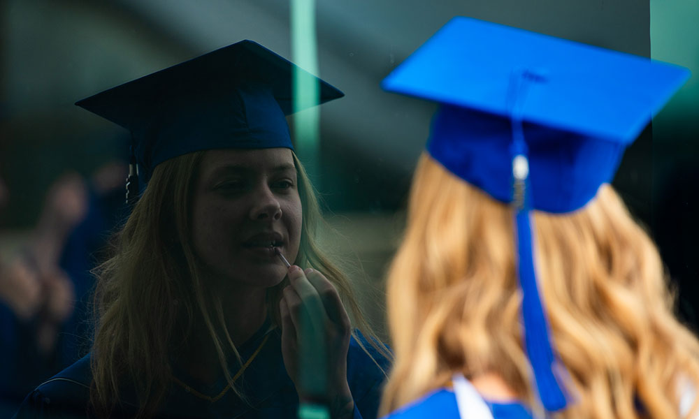 A person in a blue graduation cap and gown looking at their blurred reflection in a glass surface.