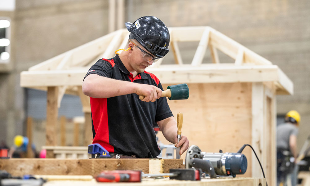 Carpenter wearing safety gear chiseling wood on a workbench with a wooden structure under construction in the background.