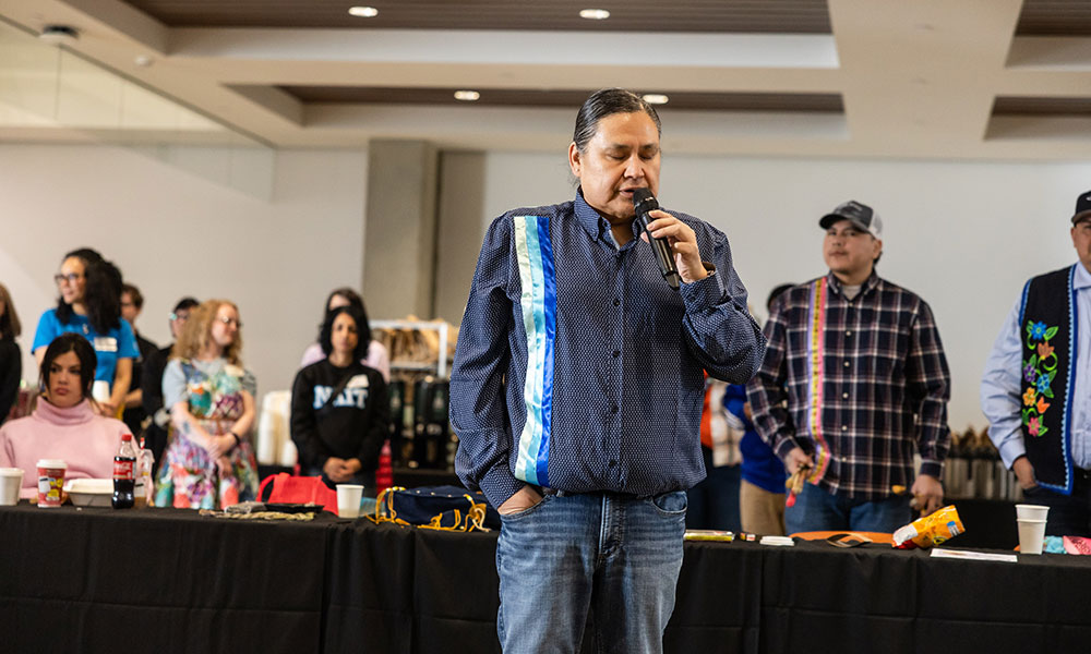 A person speaking into a microphone stands in front of a long table while a group gathers in the background at an indoor event.