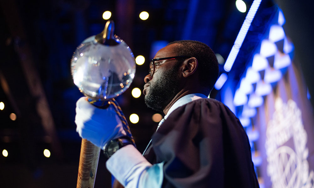 Person in formal academic attire holding a ceremonial mace with a large, clear spherical top. Background features decorative lights and geometric patterns.