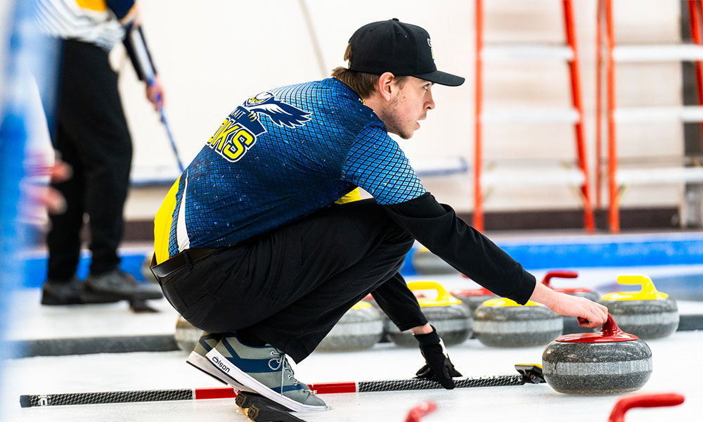 a male curler crouches and holds the handle of a red stone as he looks down the ice. he wears a blue shirt and a black ball cap.