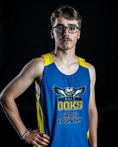 a man in a blue running tank top with the works NAIT Ooks Cross Country poses for a photo in a studio. he has medium length wavy brown hair, glasses and a moustache