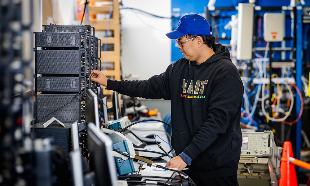 A person wearing a black NAIT hoodie works with electronic equipment on a lab bench in a technical workspace filled with servers and instruments.