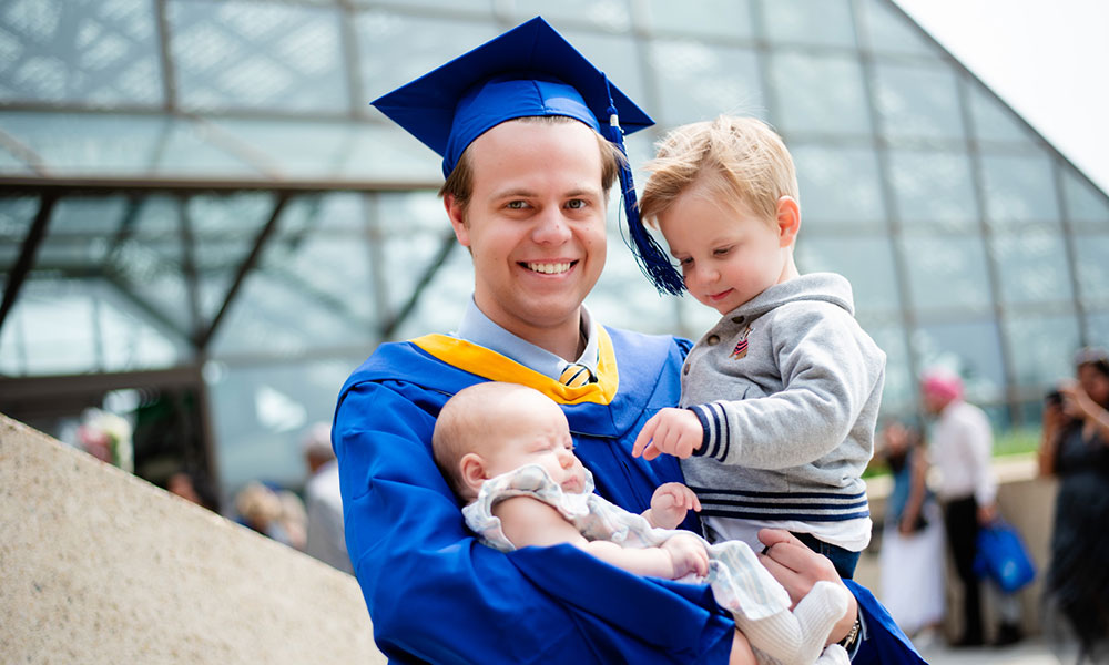 Person in a blue graduation gown and cap holding two young children, with a modern glass building and people in the background.