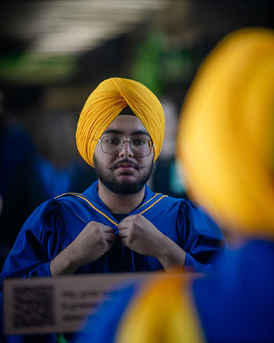 Person in a yellow turban and blue graduation gown adjusting the collar of the gown. Another individual in similar attire is partially visible from behind.