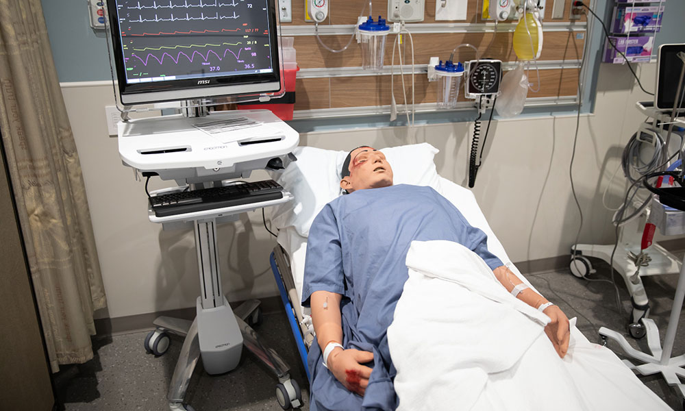 Hospital room with a mannequin patient lying in bed wearing a blue gown and covered by a white blanket. Medical equipment including an ECG monitor, IV drip, and supplies are visible around the bed.