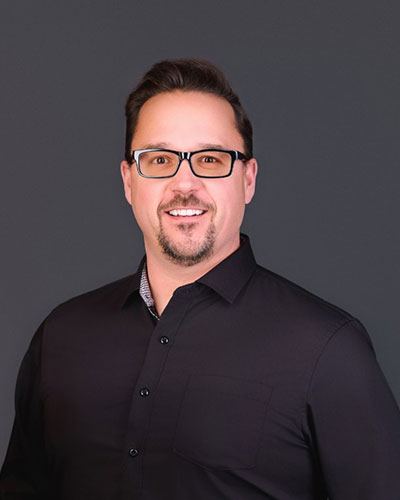 Upper‑body portrait of a person wearing a black button‑up shirt against a dark, neutral background.