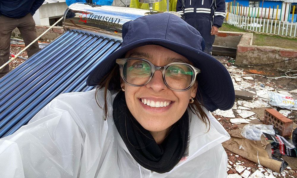 a woman wearing a blue bucket hat and glasses takes a selfie. a machine featuring many pipes side by side is over her right shoulder.