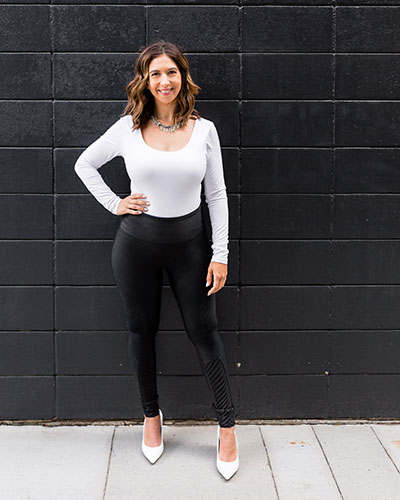 A woman standing against a black brick wall, wearing a white top, black pants, and white heels with one hand on her hip.