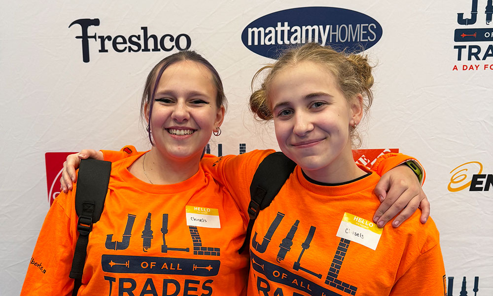 Two people are wearing bright orange “Jill of All Trades” shirts with tool graphics and name tags, standing in front of a white backdrop featuring logos like Freshco, Mattamy Homes, and Enbridge.
