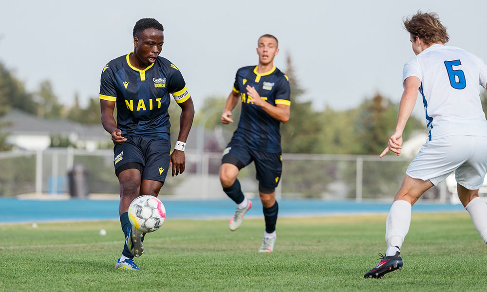 A soccer player dribbles the ball during an outdoor match as teammates and an opposing player close in on a grass field.