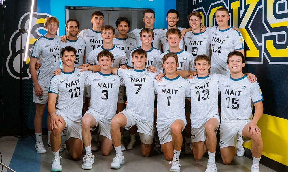 A group of uniformed NAIT volleyball players pose together for a team photo in front of a brightly colored athletic backdrop.