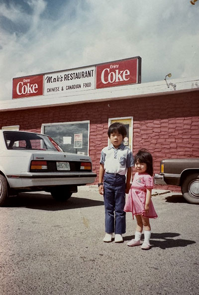 Two children standing in front of Mak's Restaurant, which has signage reading 'Enjoy Coke' and 'Chinese & Canadian Food.' Two cars are parked near the entrance.