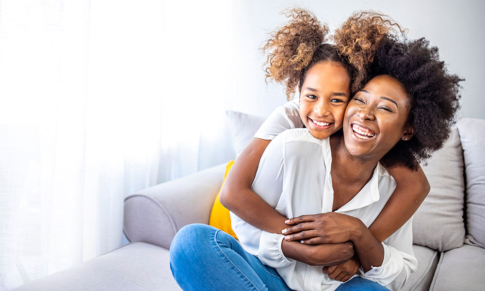 Adult and child hugging on a couch with sunlight coming through a window.