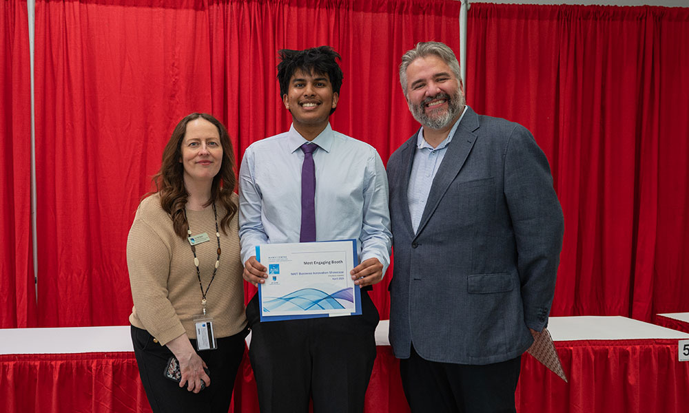 a photo of a man in a blue blazer, a man in a shirt with a purple tie, and a woman in a brown sweater. the man with the tie, in the middle, holds a certificate.