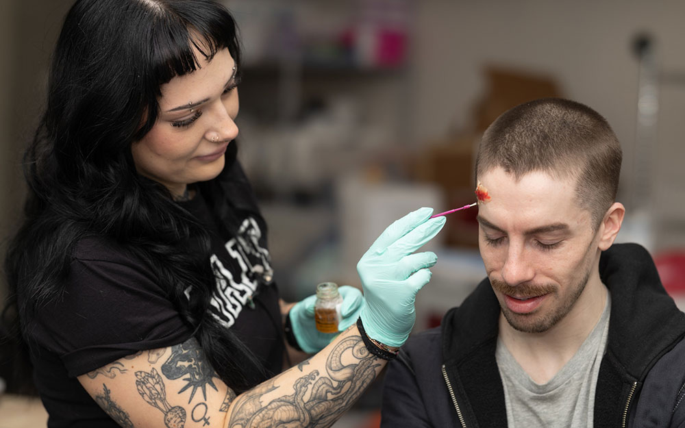 Instructor wearing gloves applies moulage makeup to a seated man’s forehead during a practical training session.